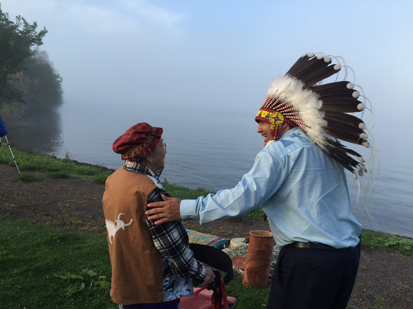 Perry Bellegarde in headdress greeting Elder Irene Stevens at a foggy Lake Superior shore